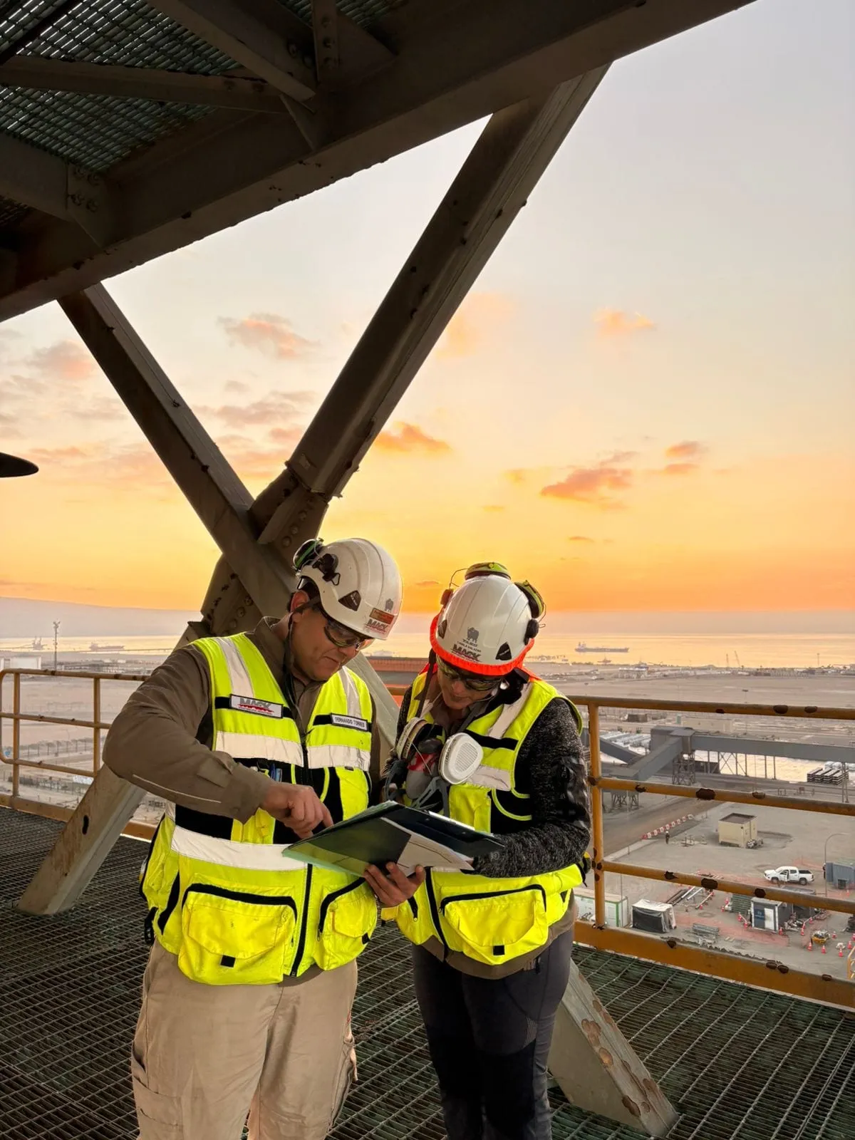 Dos trabajadores de MACK revisan documentos en una pasarela al atardecer